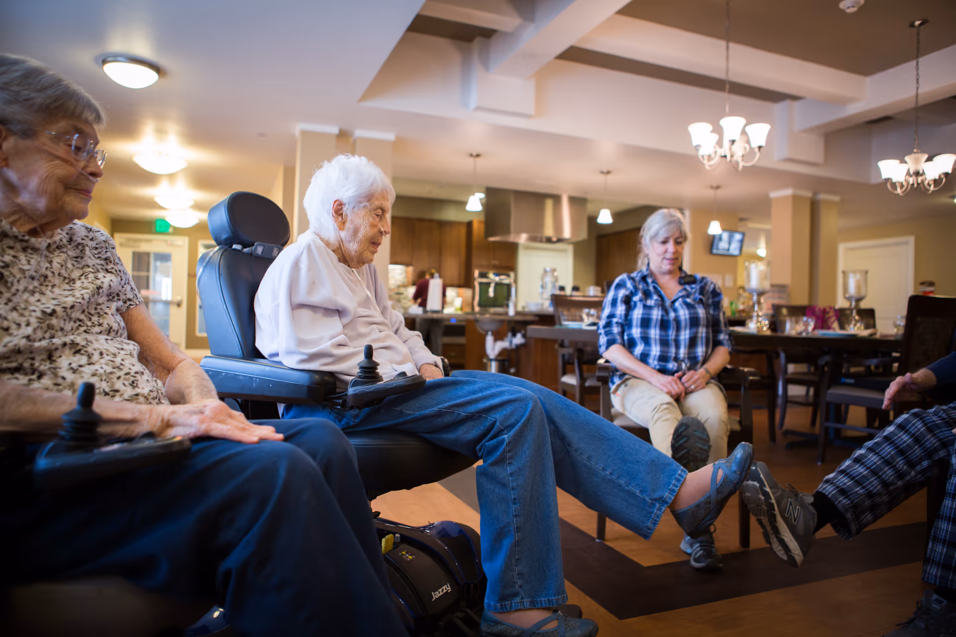 Several elderly residents and a staff member sit in a communal dining area with a kitchen visible in the background.