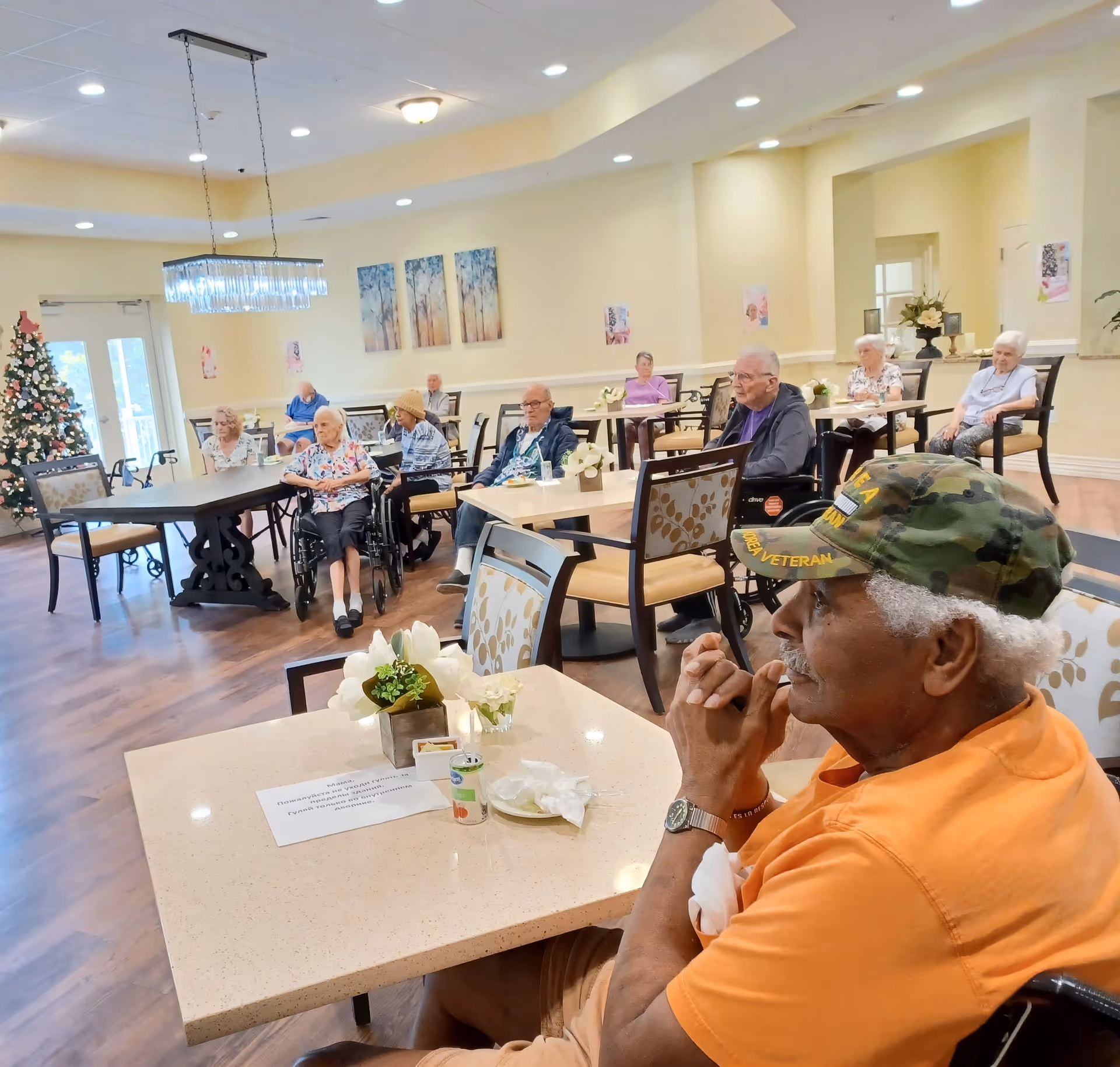 A group of elderly residents sitting in a spacious, well-lit common area with tables and chairs. Some residents are in wheelchairs, and the room features wooden flooring, a decorated Christmas tree, and wall art. The atmosphere appears calm and communal.