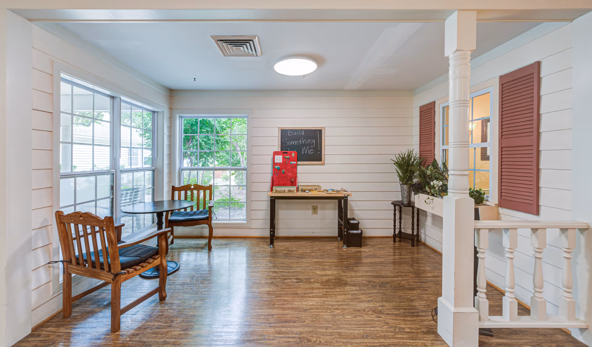 A bright room with large windows showing greenery outside, two wooden chairs with cushions around a small round table on the left, a table against the wall with a red activity board and a chalkboard above it that says 'Build Something Me!', and a small table with potted plants near a window with red shutters on the right. The floor is wooden and the walls are white with horizontal paneling.