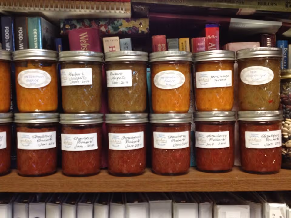 Two rows of homemade jam jars with handwritten labels on a wooden shelf. The top row contains jars labeled with flavors like marmalade sauce, rhubarb jalapeno, and persimmon spread. The bottom row contains jars labeled strawberry rhubarb jam. Behind the jars are books on a bookshelf.