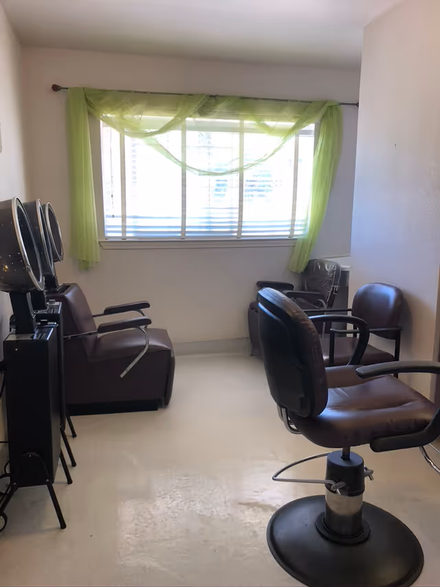 A small hair salon area with two brown salon chairs and two hair dryer stations against the left wall. A window with green sheer curtains lets in natural light, and the floor is light-colored.