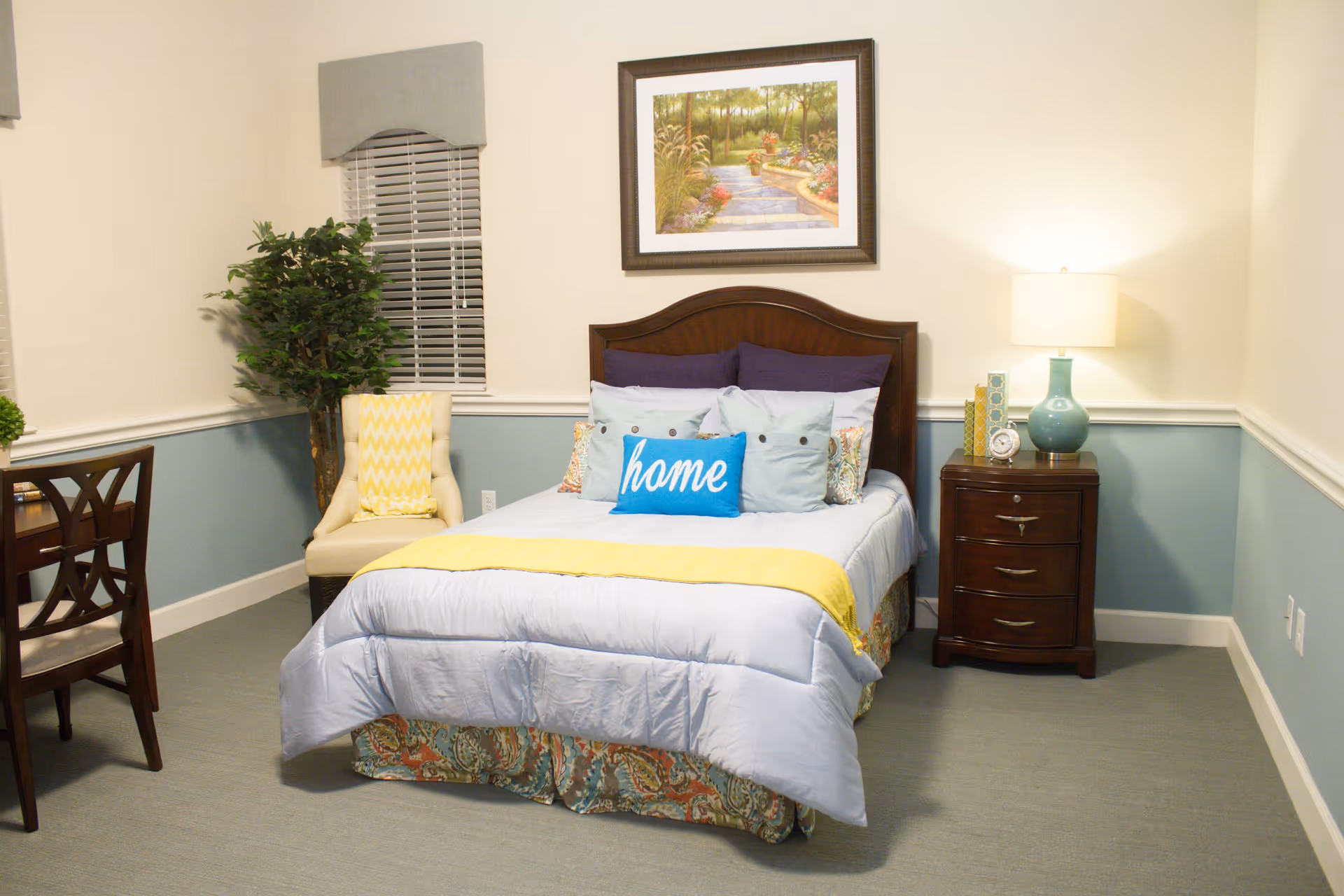 A cozy bedroom with a neatly made bed featuring multiple pillows including one with the word 'home'. The room has light blue walls with white trim, a wooden nightstand with a lamp, a framed painting above the bed, a cushioned chair with a yellow throw, and a wooden desk with a chair.