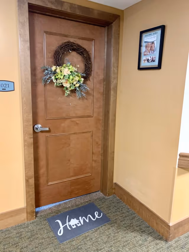 Closed wooden apartment door in a facility hallway decorated with a floral wreath and a 'Home' doormat.