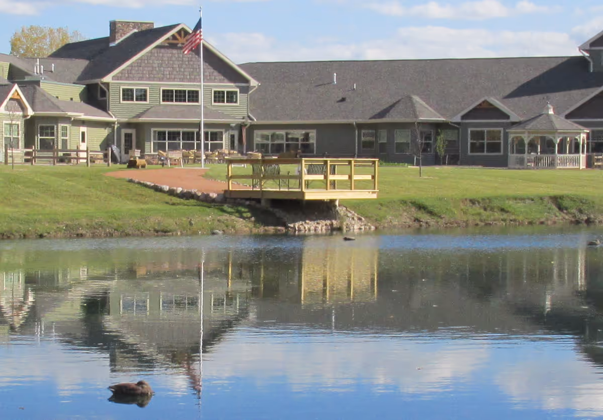 A lakeside assisted living building with a gazebo, dock, American flag, and lawn reflected in a pond.