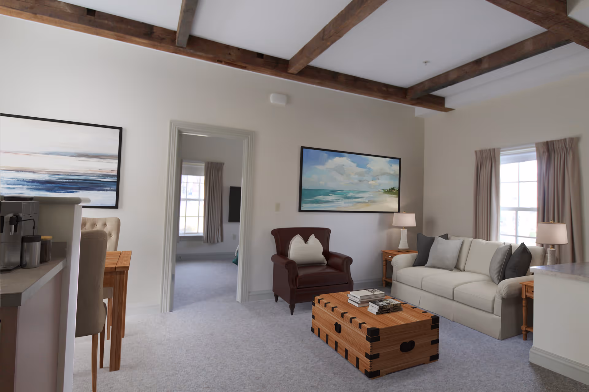 A cozy living room with a white sofa adorned with gray and white pillows, a brown leather armchair with a white pillow, and a wooden chest-style coffee table with books on top. The room has beige walls, two windows with beige curtains, two side tables with lamps, and two framed paintings on the walls. The ceiling features exposed wooden beams. A doorway leads to another room with a window and curtains.