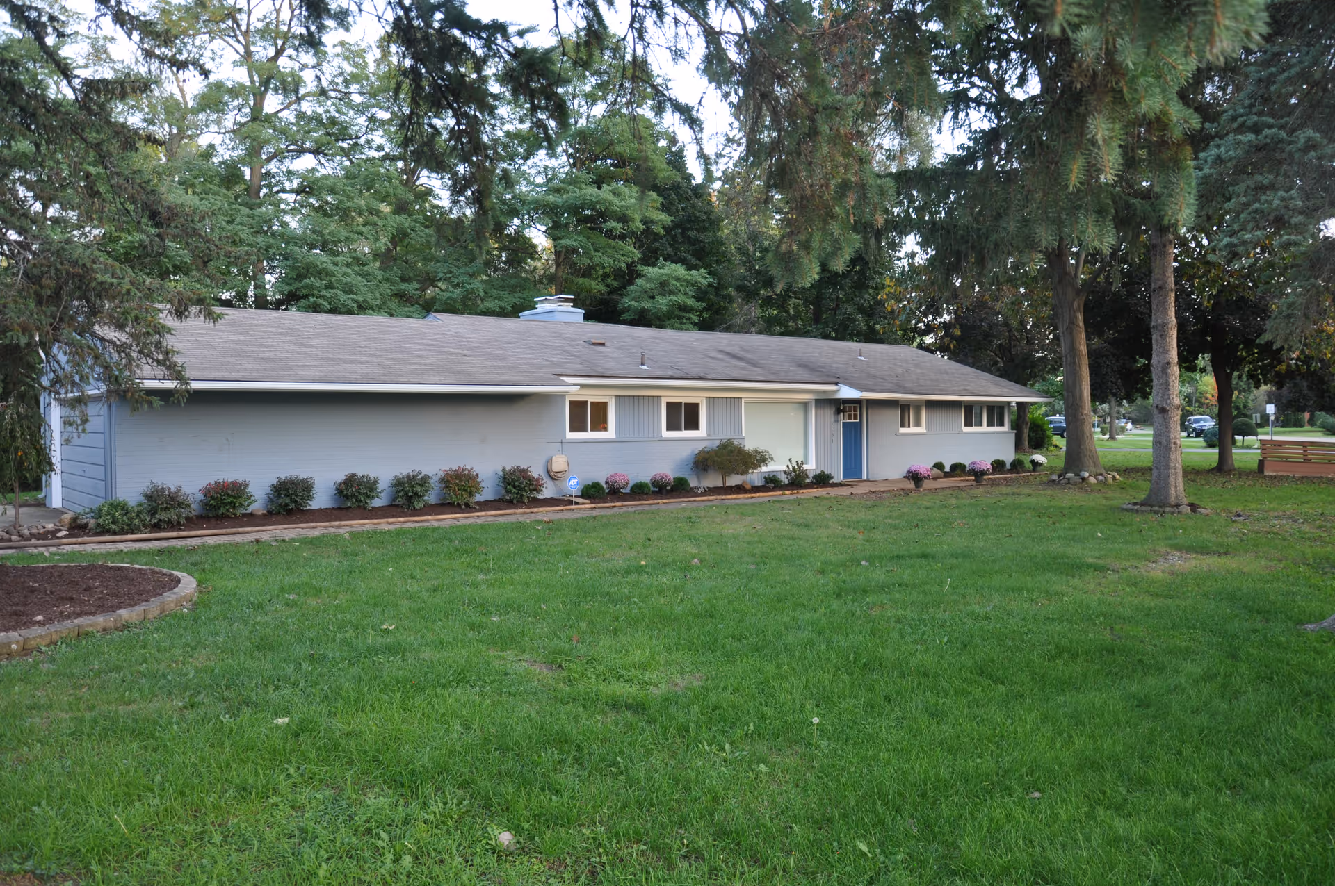 Single-story building with a gray exterior and a blue door, surrounded by green grass, trees, and small shrubs along the front. The building is set in a park-like environment with a bench visible in the background.