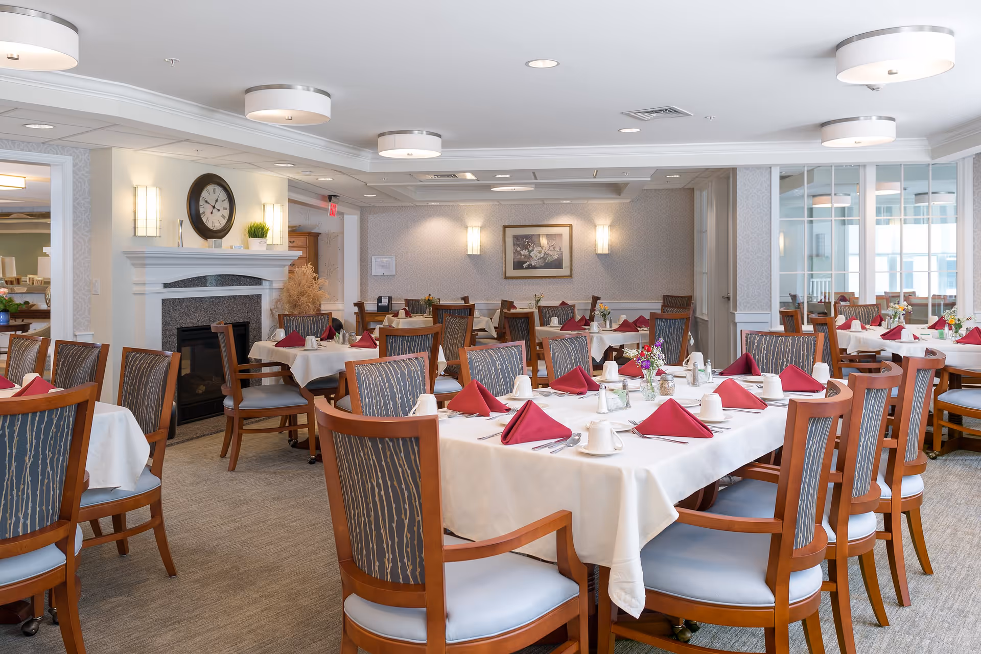 A bright and elegant dining room with multiple tables covered in white tablecloths, each set with red folded napkins, white cups, and silverware. The room features wooden chairs with patterned upholstery, a fireplace with a clock above it, soft wall lighting, and a framed floral painting on the far wall. Large windows and glass panels allow natural light to fill the space.