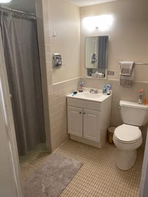 A small bathroom with beige tiled walls and floor. There is a white toilet with a towel hanging on a rack above it, a white vanity with a sink and a mirror above it, and a shower area with a gray curtain. Various toiletries are placed on the sink and toilet tank.