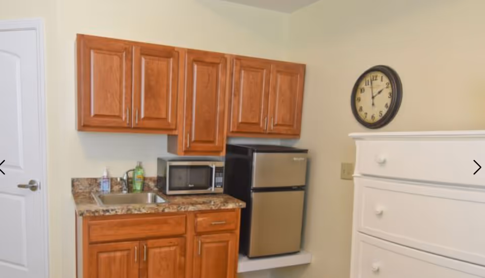 Small kitchenette area with wooden cabinets, a granite countertop with a sink, a microwave, and a compact stainless steel refrigerator. A white chest of drawers is visible to the right, and a round wall clock hangs above it on a beige wall.
