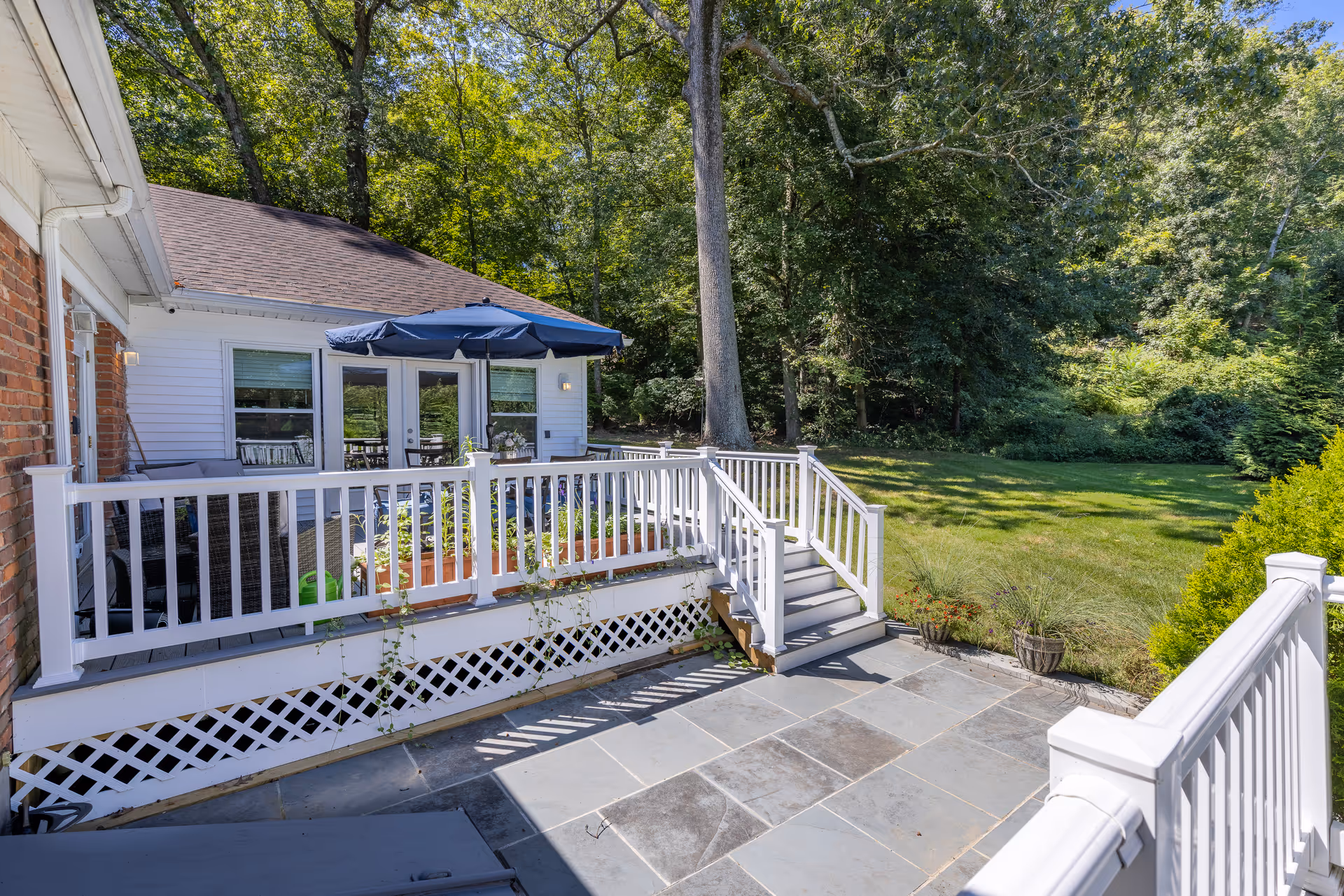Outdoor patio area with stone tile flooring, white railing, and steps leading up to a deck with a blue umbrella and outdoor seating. The patio is adjacent to a white house with large windows and surrounded by green trees and grass.