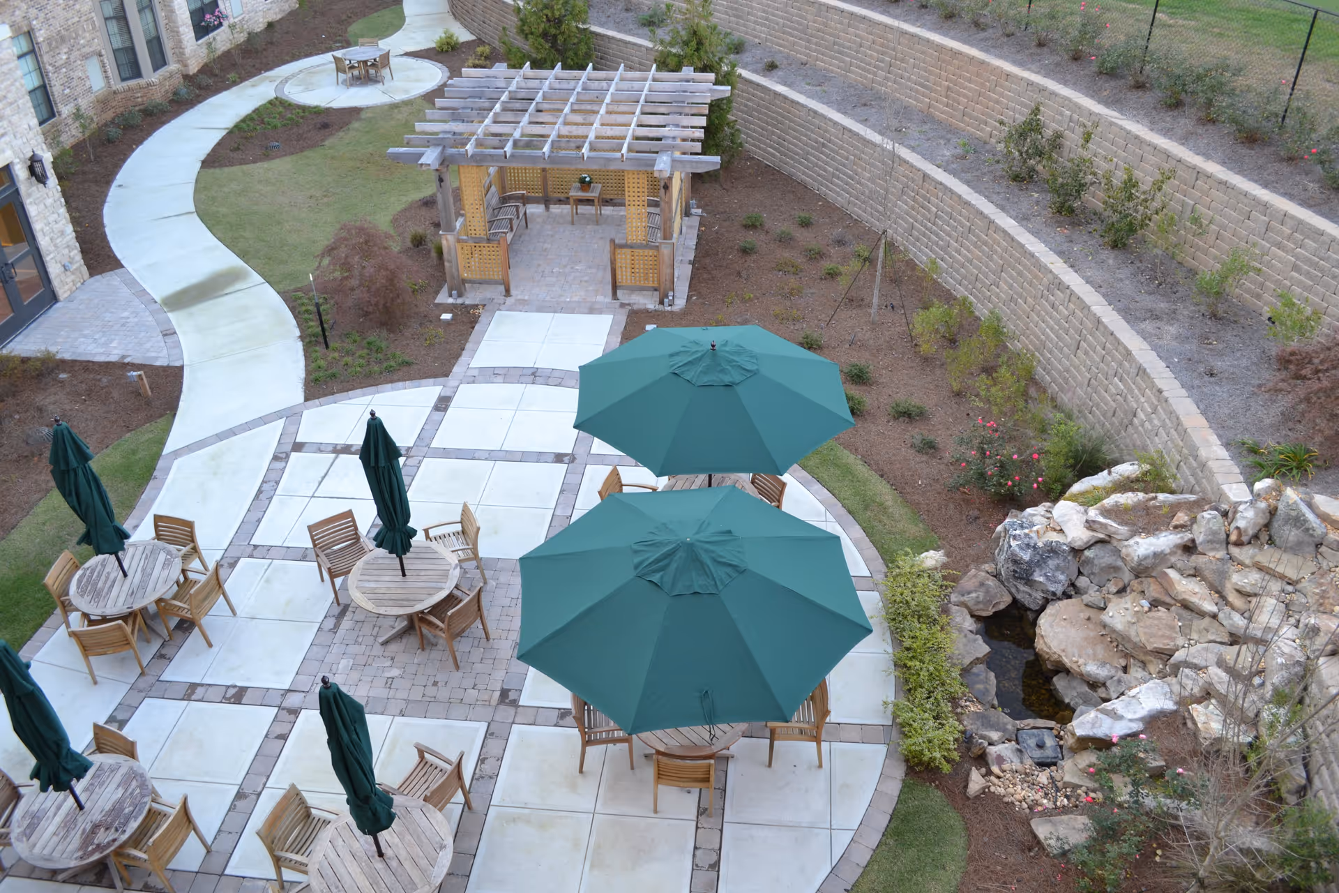 Outdoor patio area with several round wooden tables and chairs, some tables shaded by large green umbrellas. A curved concrete walkway leads to a wooden pergola with seating underneath. There is a stone retaining wall with plants and a small rock water feature on the right side.