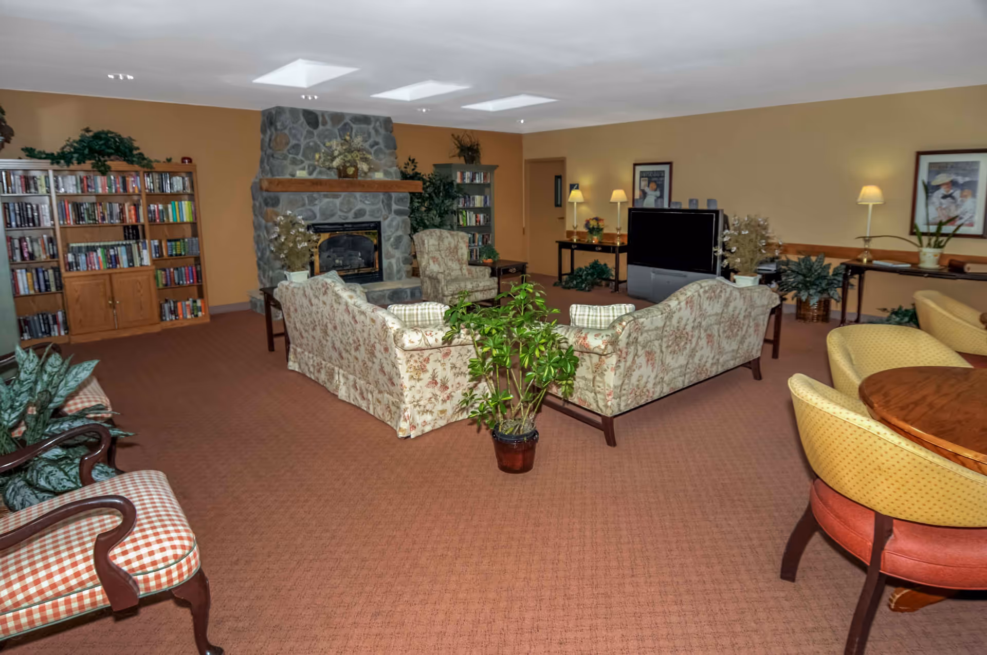 Communal living room with floral sofas arranged around a stone fireplace, bookshelves, a TV, plants, and additional seating areas.