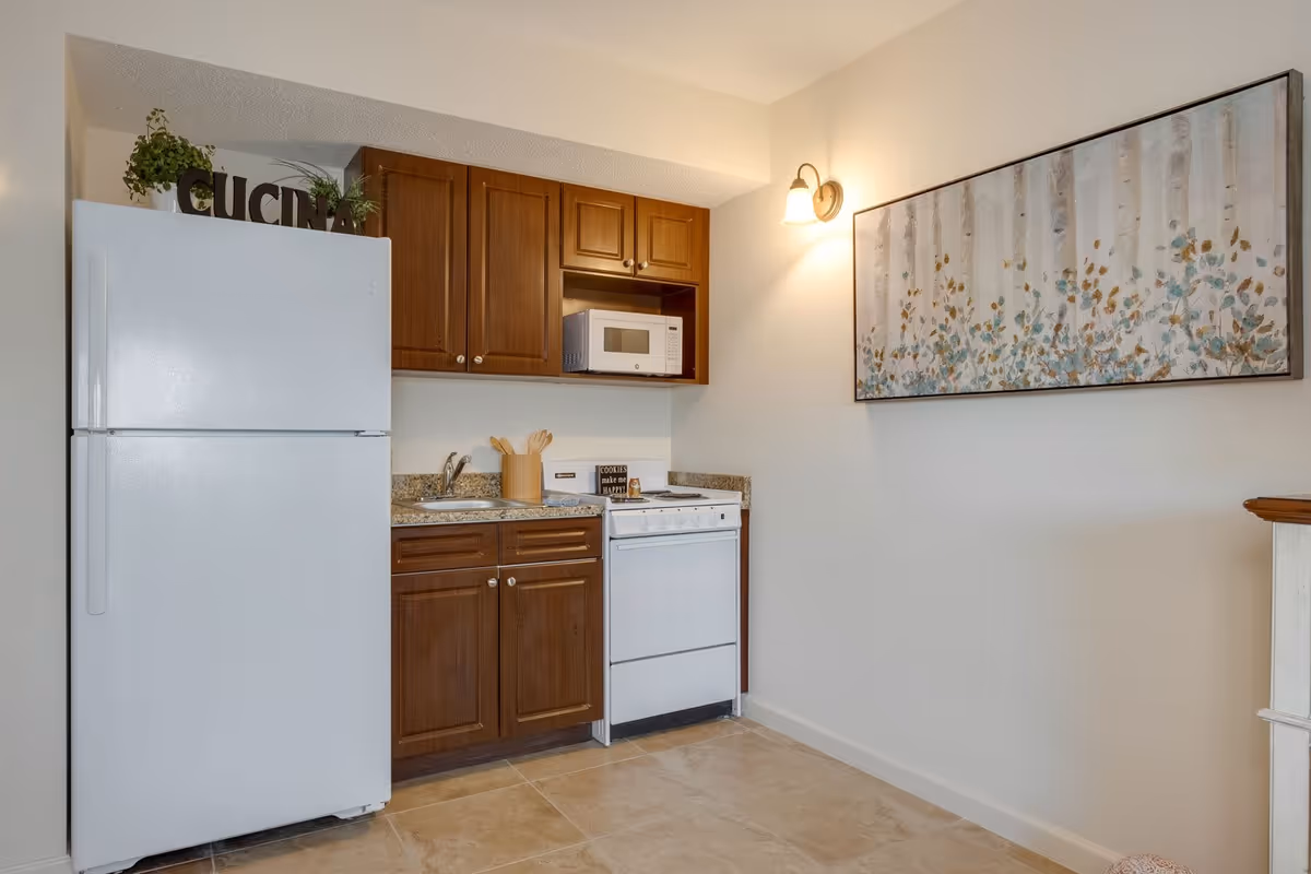 A small kitchen area with a white refrigerator, wooden cabinets, a microwave, a stove, and a sink. There are decorative plants and a sign that says 'CUCINA' on top of the refrigerator. A wall-mounted light fixture illuminates a framed artwork featuring a floral design on the adjacent wall.