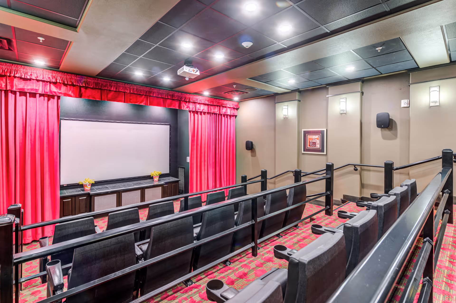 Interior view of a small theater room with red curtains framing a large white projection screen. The room has two rows of black cushioned seats with cup holders, red patterned carpet, beige walls with wall-mounted lights, and a ceiling-mounted projector.