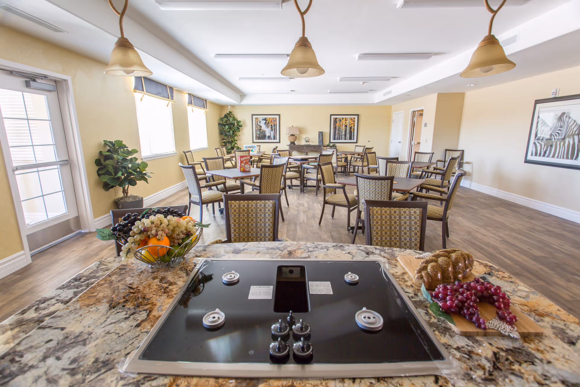 Bright communal dining room with multiple tables and chairs viewed from a kitchen island with a stovetop and fruit bowls.