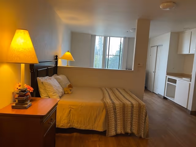 A cozy bedroom with a bed covered in a beige blanket and a knitted throw, two pillows, and a small teddy bear. A wooden nightstand with a lamp and a stack of books is beside the bed. The room has wooden flooring, a large window letting in natural light, and a partial view of a kitchenette area with white cabinets and a countertop.