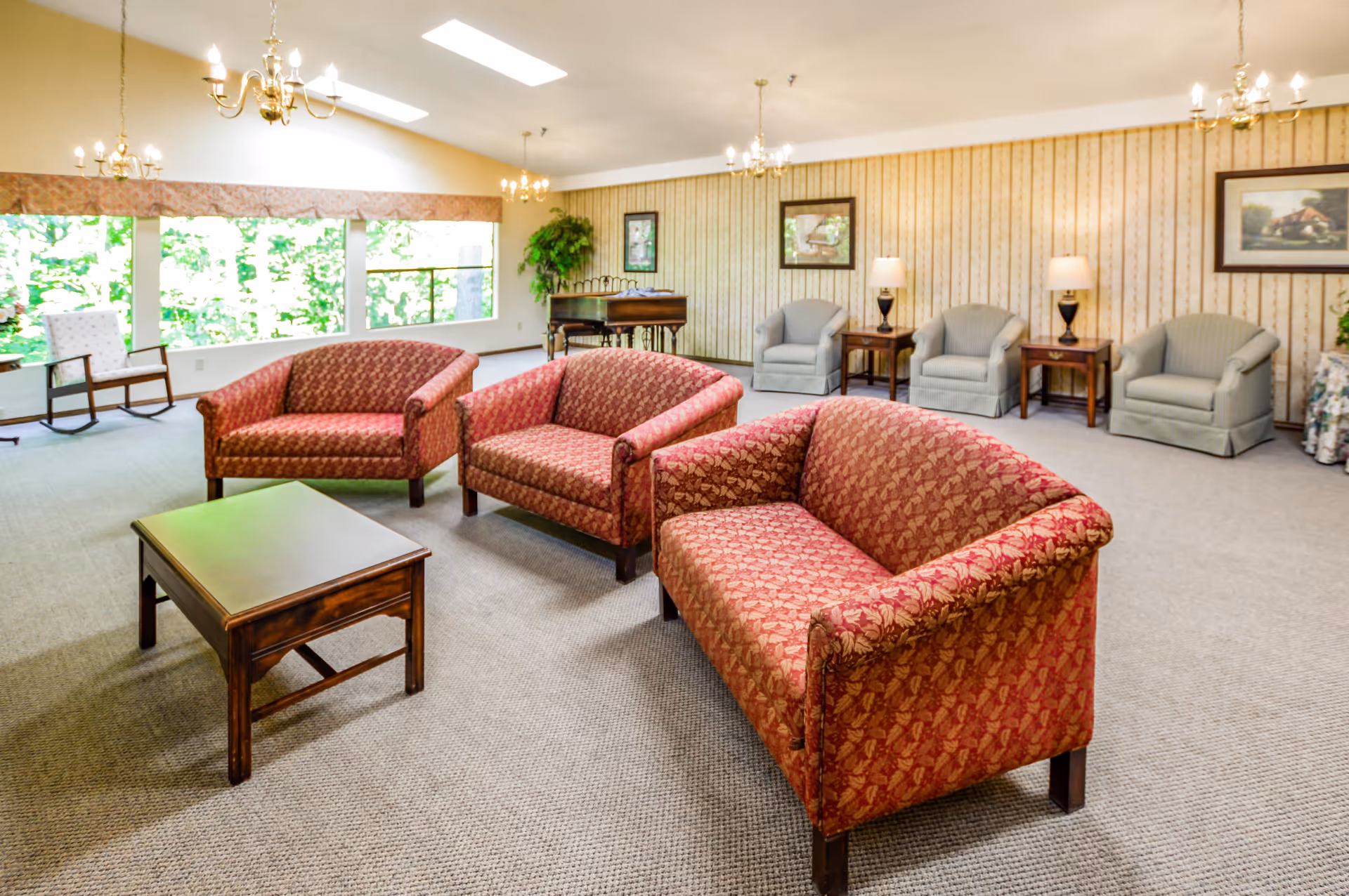 Spacious assisted-living common room with patterned red sofas, armchairs, a coffee table, chandeliers, and large windows overlooking greenery.