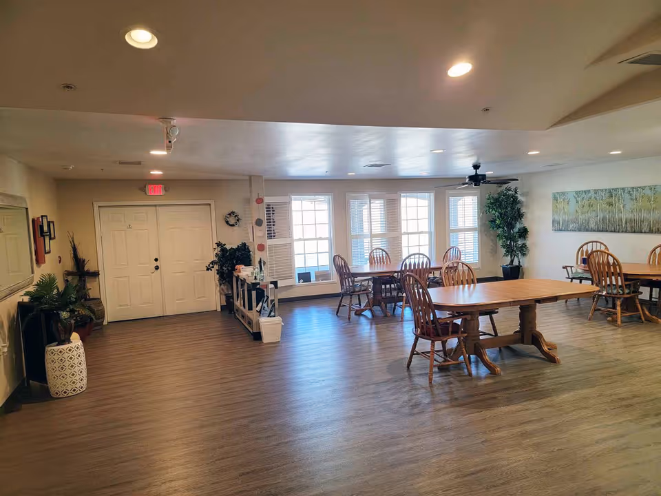Spacious communal dining room with wooden tables and chairs, large windows, and potted plants.