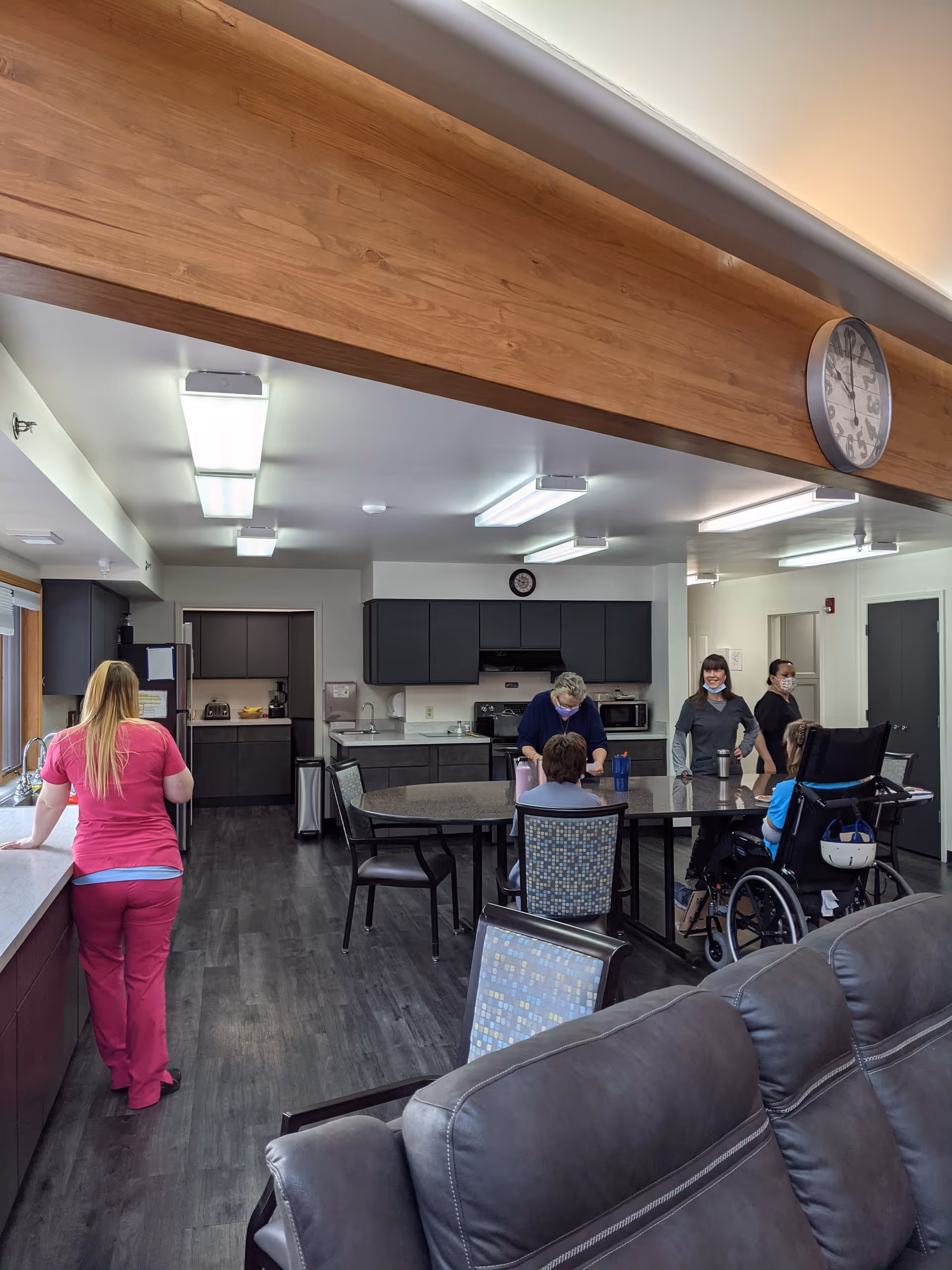 Interior view of a rehabilitation facility kitchen and common area with several people, including a person in a wheelchair, gathered around a table. A staff member in pink scrubs stands near the counter. The room has dark flooring, gray cabinets, and bright overhead lighting. A large clock is mounted on a wooden beam above.