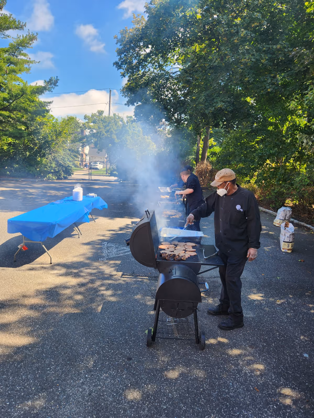 Two people cooking on outdoor grills in a parking lot area surrounded by trees. One person is wearing a mask and a cap, tending to food on the grill, while the other is further back also grilling. A table covered with a blue tablecloth is set up nearby with some items on it. The scene is sunny with a clear blue sky and some scattered clouds.