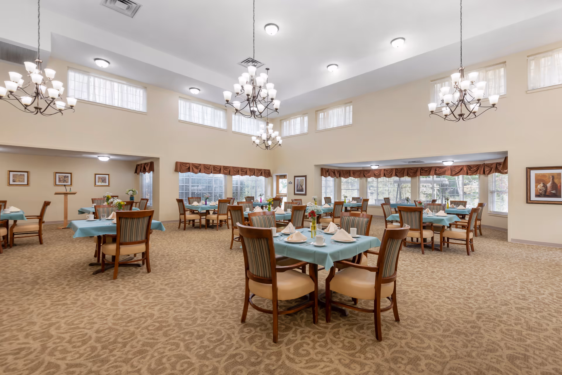 Spacious dining room with multiple round tables set with teal tablecloths and place settings, wooden chairs, chandeliers, and large windows.