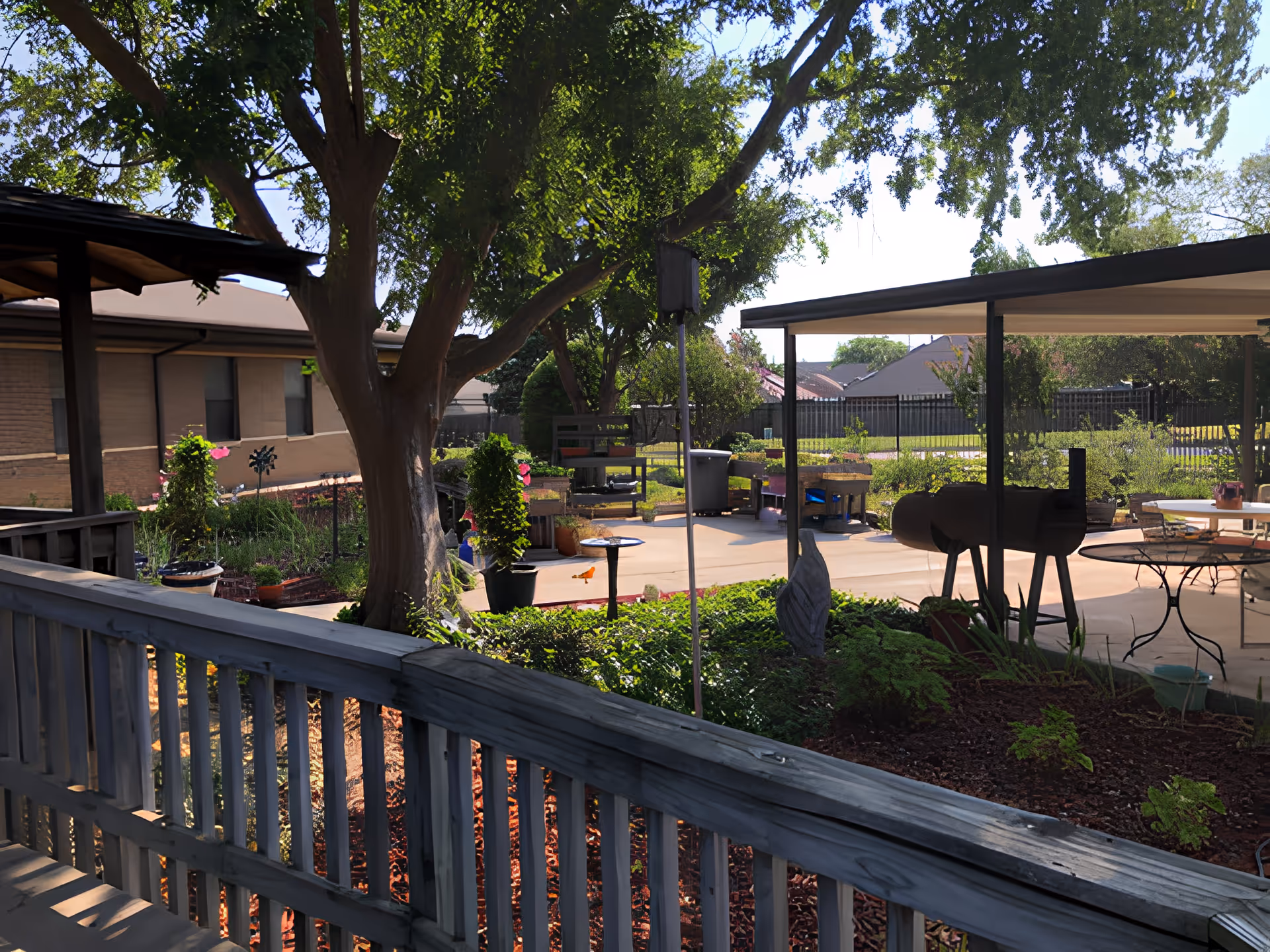 Shaded outdoor courtyard with a large tree, raised garden beds, a covered patio area with tables and a grill, and a wooden railing in the foreground.
