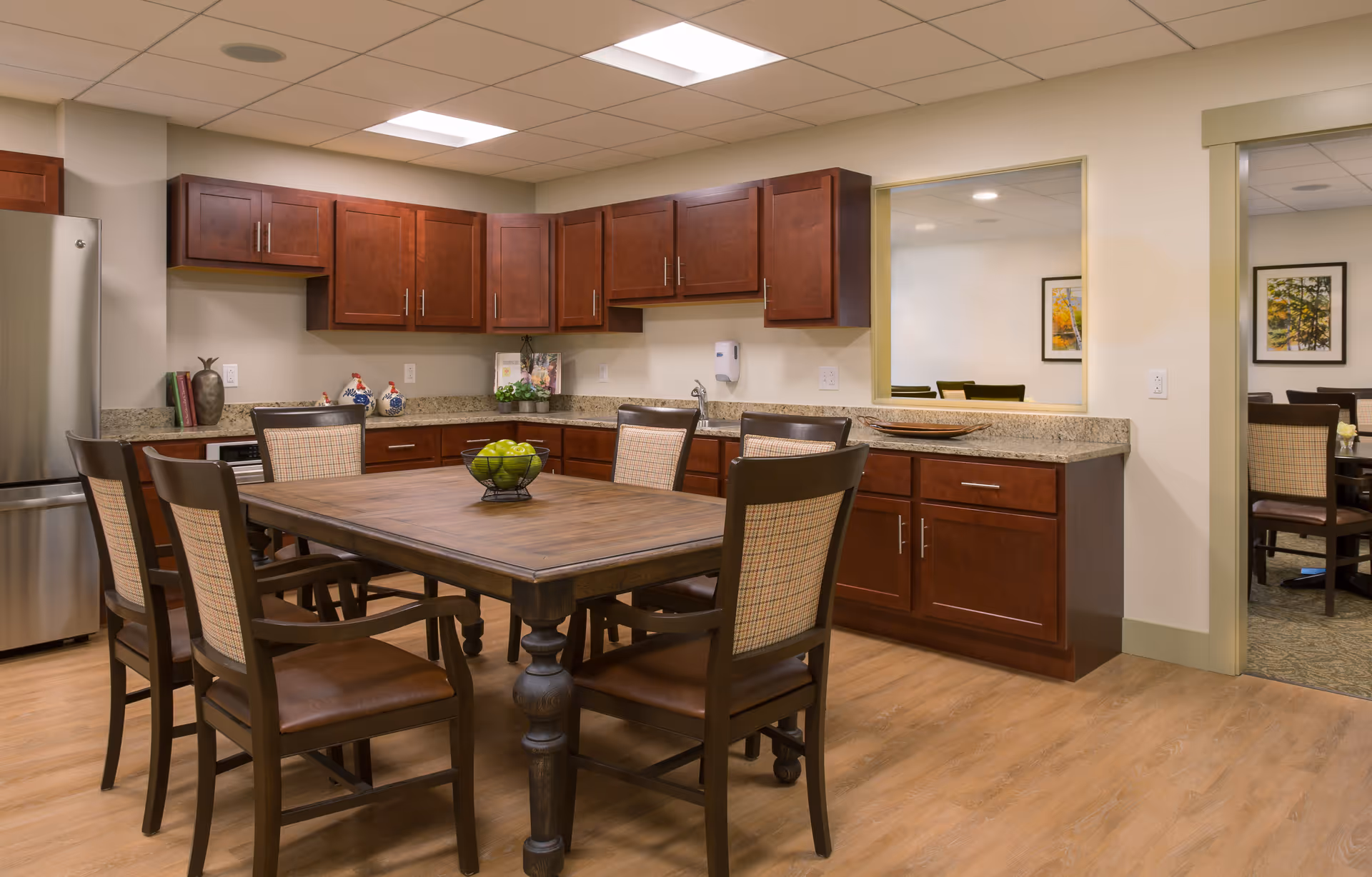 A kitchen and dining area with wooden cabinets, granite countertops, and a wooden dining table surrounded by six chairs. The kitchen features a stainless steel refrigerator, decorative items on the counter, and a pass-through window looking into another room with framed artwork on the walls.