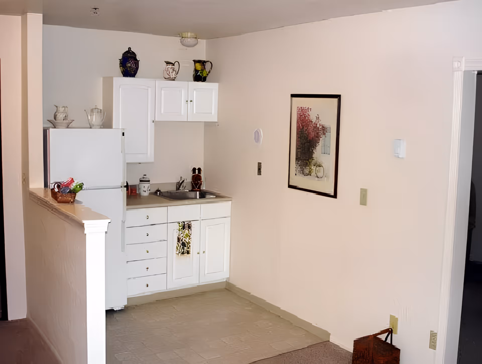 Small kitchen area with white cabinets, a white refrigerator, a sink, and a countertop. Decorative pitchers and a basket with fruit are placed on top of the cabinets and refrigerator. A framed picture hangs on the adjacent wall.
