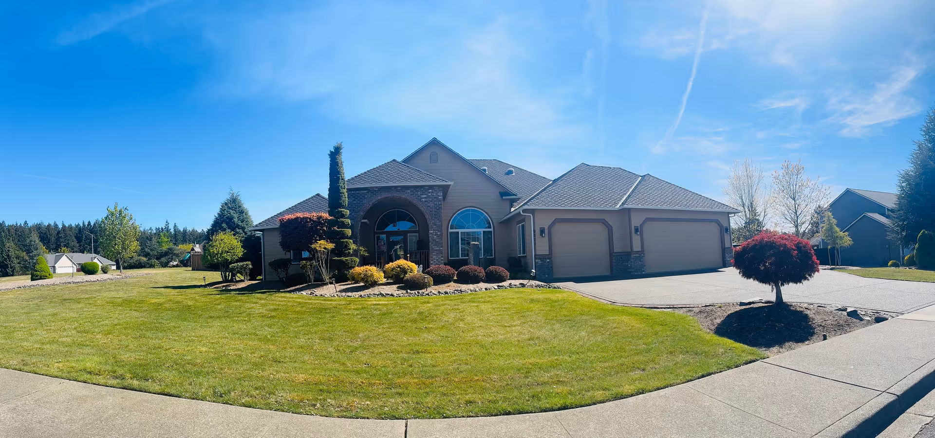 A single-story residential building with a large front lawn, well-maintained shrubs, and a clear blue sky overhead. The house features a three-car garage and a prominent arched entrance with large windows.