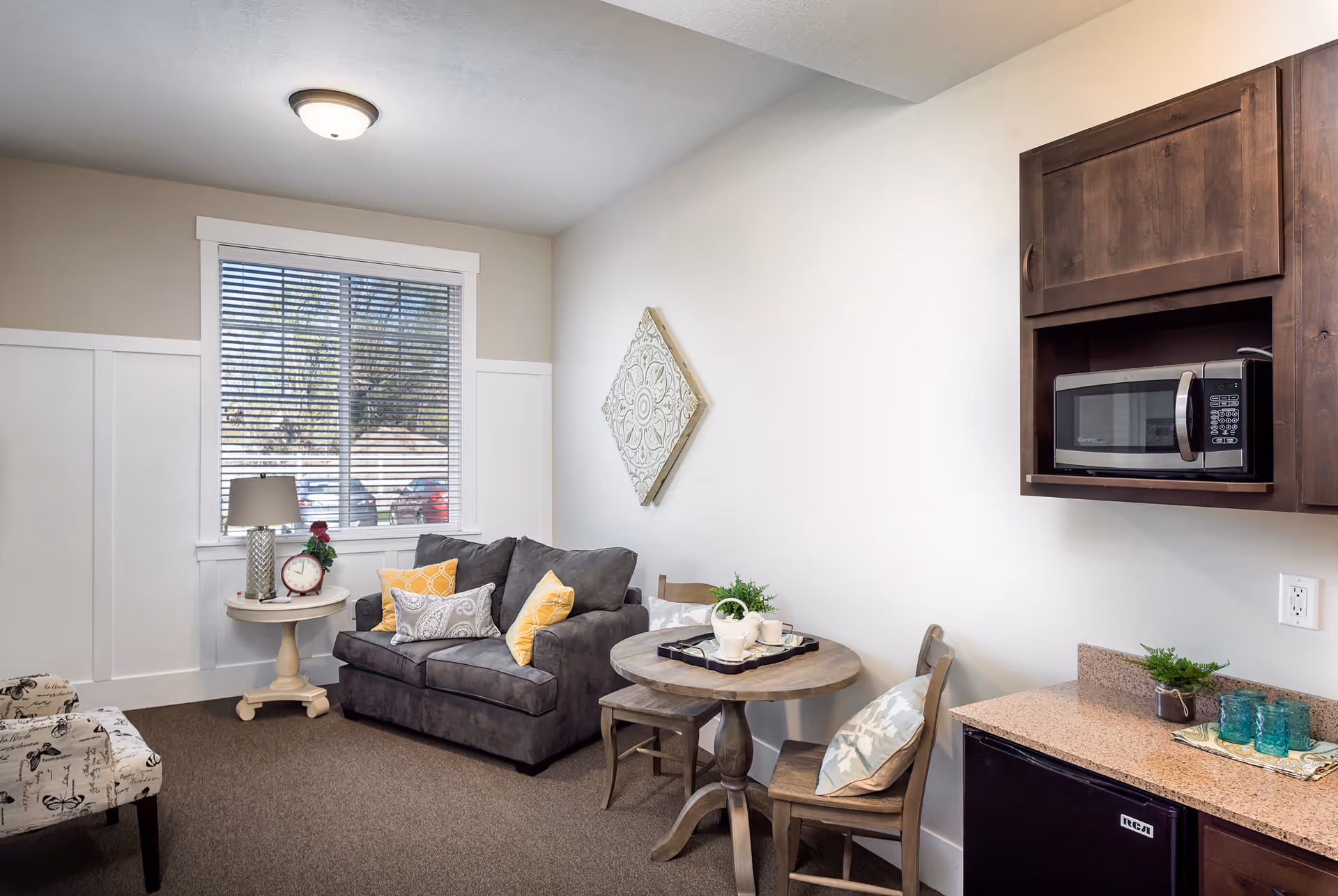 A cozy living area with a gray loveseat adorned with yellow and patterned pillows, a small round wooden table with two chairs, a side table with a lamp and clock, a window with blinds, and a kitchenette area with a microwave and mini fridge.