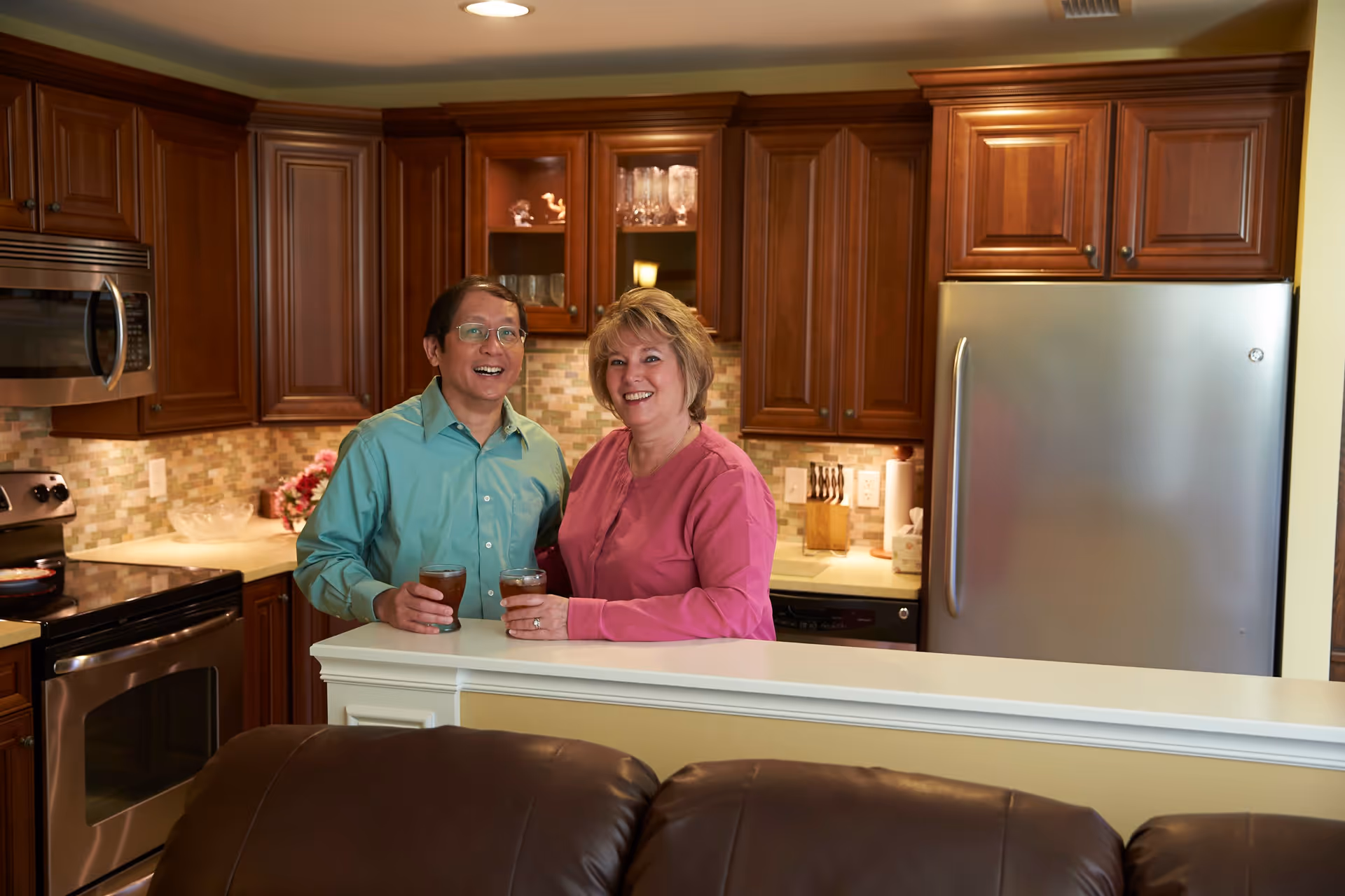A man and a woman standing together in a kitchen, smiling and holding glasses of iced tea. The kitchen features wooden cabinets, a stainless steel refrigerator, microwave, and stove, with a tiled backsplash and a countertop separating the kitchen from the living area.