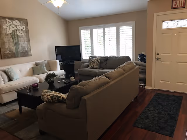 A cozy living room with two beige sofas and one gray sofa arranged around a dark wooden coffee table. A flat-screen TV is placed on a stand near a window with white shutters. The room has wooden flooring, a dark rug near the entrance door, and a ceiling fan with a light fixture. A floral painting hangs on the wall above one of the sofas.