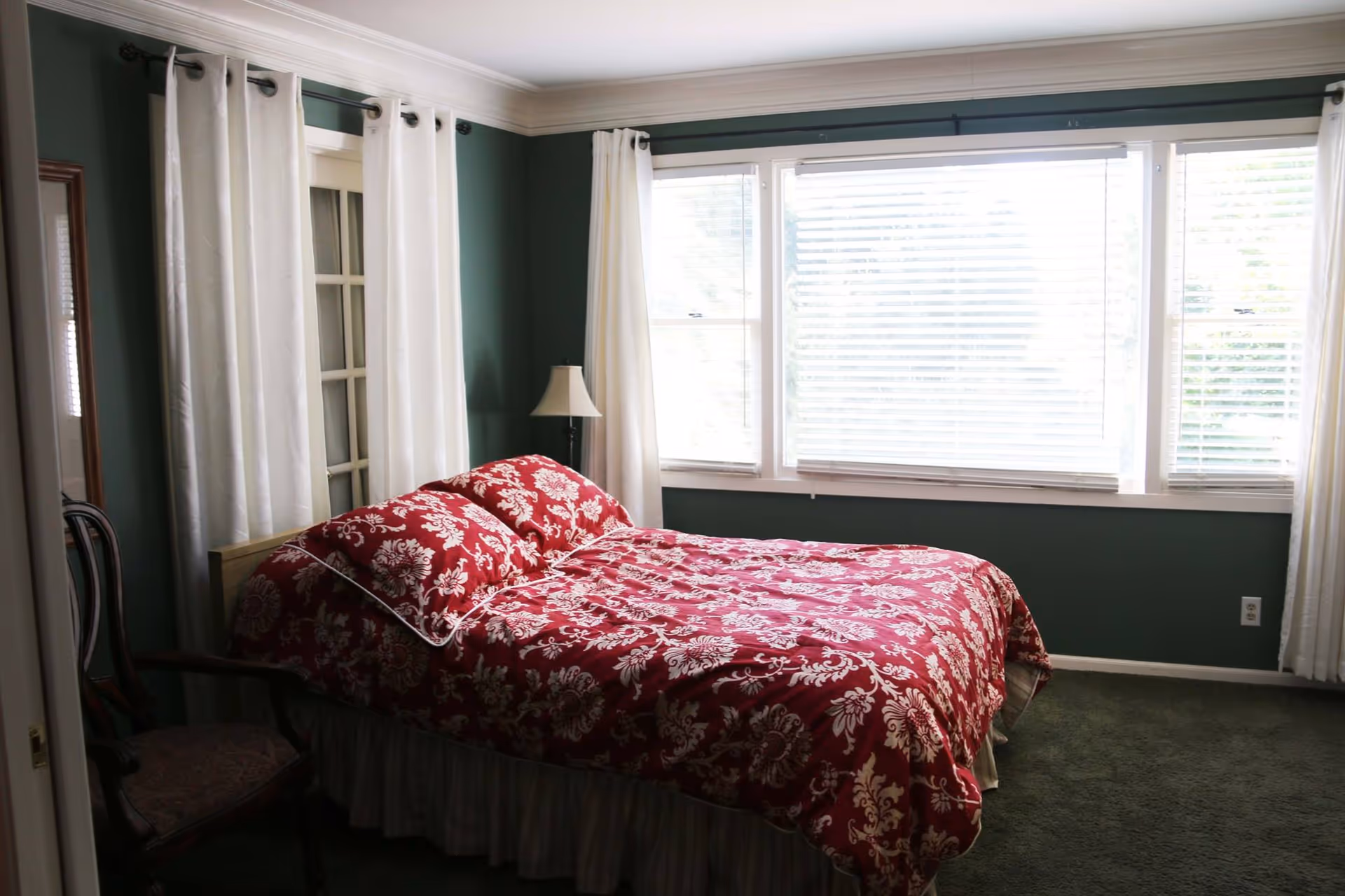 A bedroom with a bed covered in a red and white floral patterned comforter and matching pillows. The room has green walls, white curtains on the windows, a wooden chair with a cushioned seat, and a floor lamp next to the bed. Large windows with blinds allow natural light into the room.