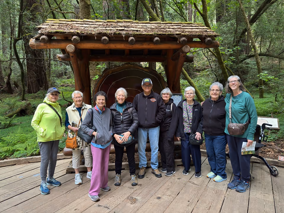A group of nine older adults standing on a wooden platform in a forested area, posing in front of a large wooden structure with a cross-section of a tree trunk displayed behind them. They are dressed in casual outdoor clothing and appear to be enjoying a nature outing.