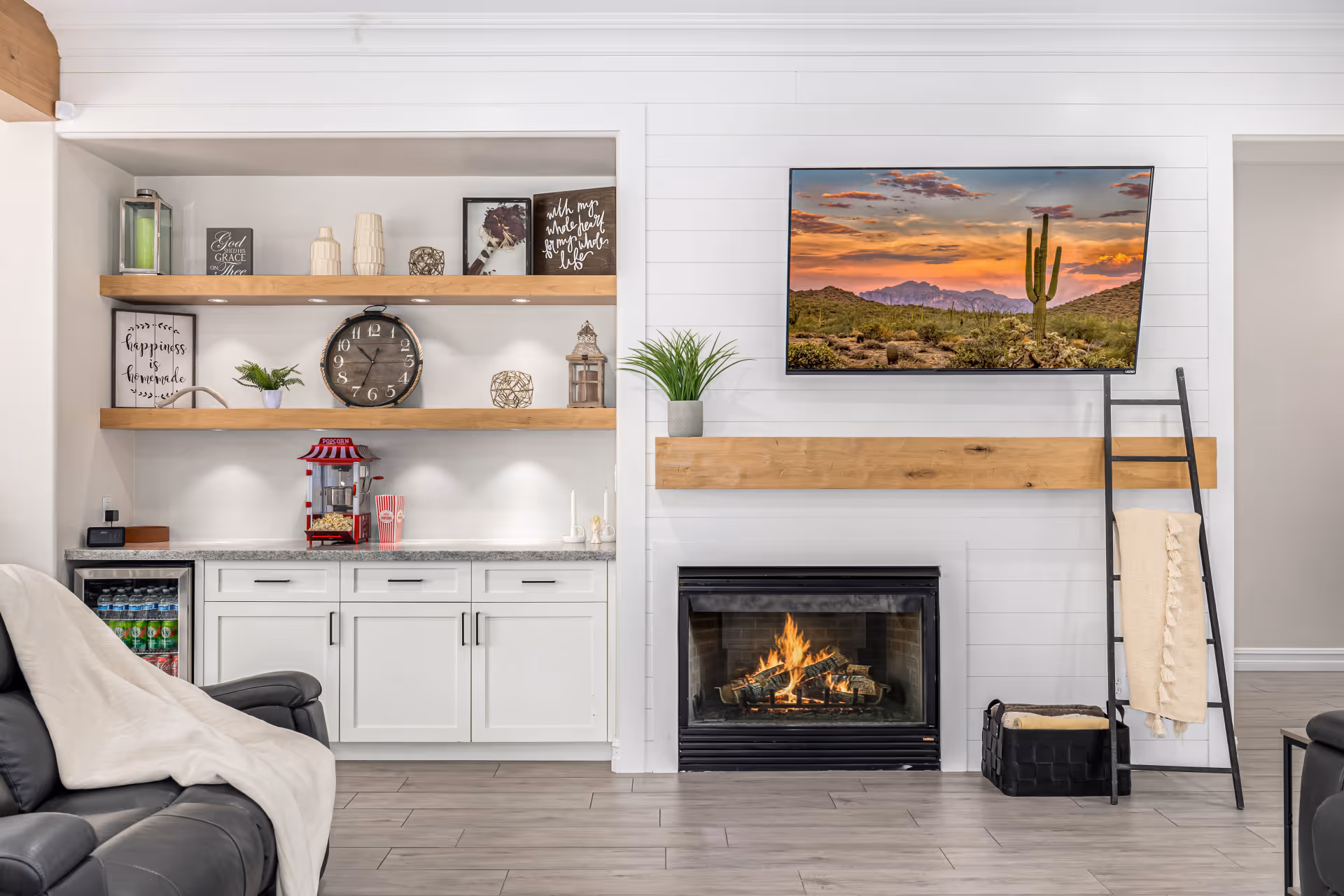 A cozy living room area with a lit fireplace beneath a mounted flat-screen TV displaying a desert sunset scene. To the left, there are white cabinets with a granite countertop, open wooden shelves decorated with various items including a clock, framed quotes, and a popcorn machine. A black leather chair with a light-colored blanket draped over it is partially visible in the foreground. A black ladder with a beige throw blanket leans against the wall next to a basket.