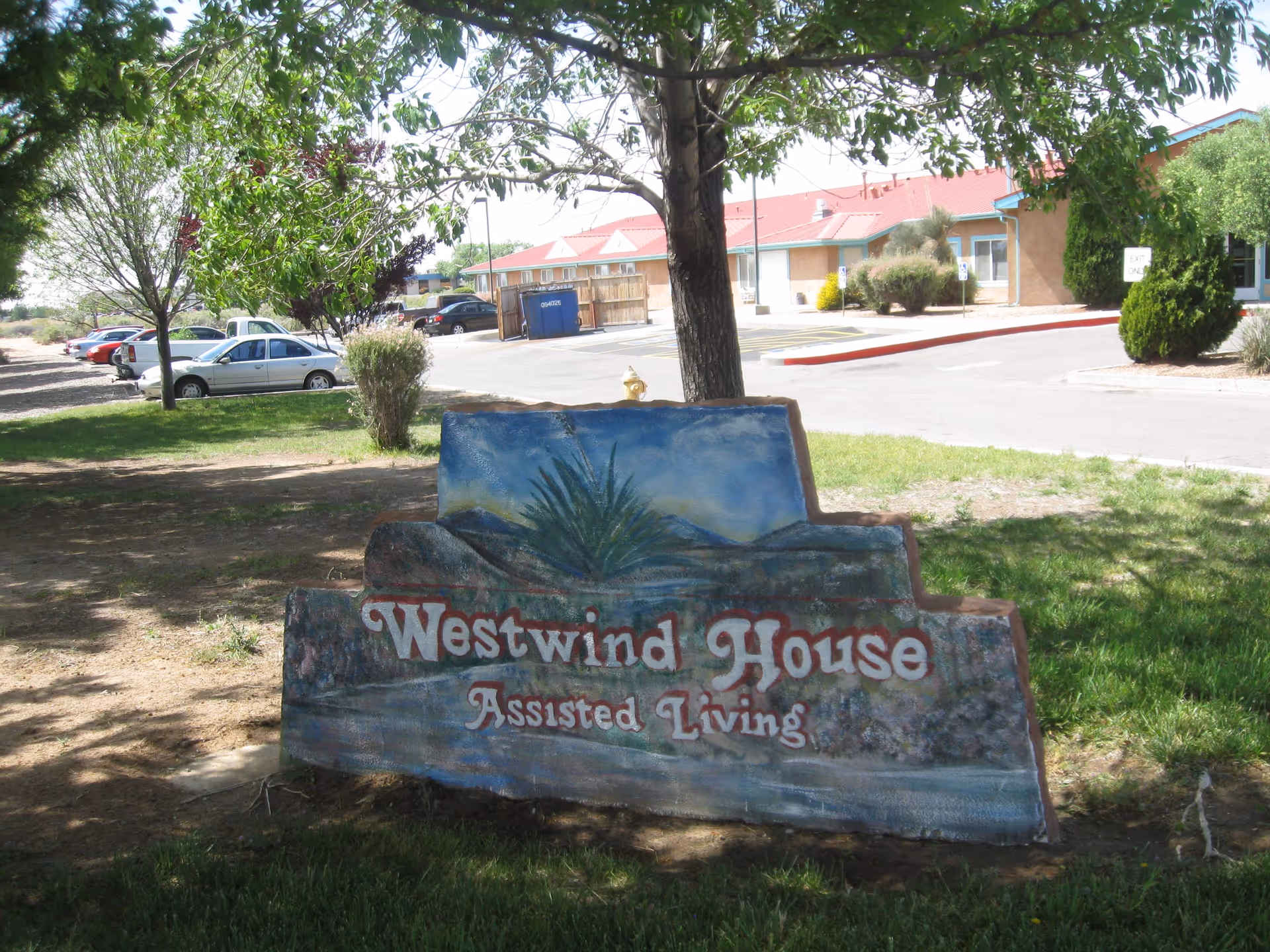 Outdoor view of a painted stone sign that reads 'Westwind House Assisted Living' with a background of trees, grass, parked cars, and a building with a red roof.