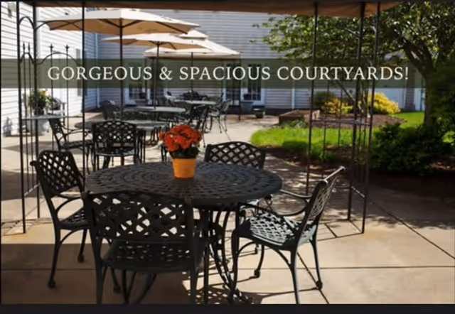 Outdoor courtyard area with multiple black metal tables and chairs, some tables have umbrellas. A potted plant with red flowers is placed on the nearest table. The courtyard is surrounded by greenery and building walls.