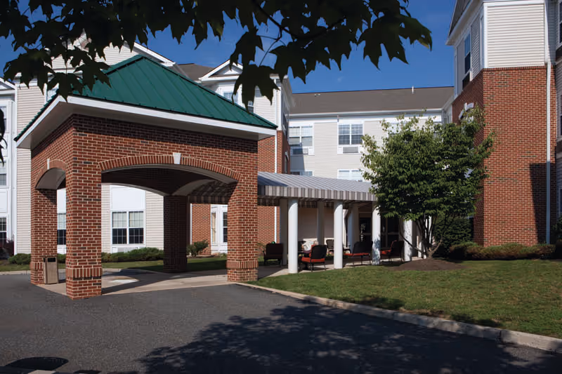 Exterior view of a senior living facility with a covered entrance supported by brick pillars and a green metal roof. The building is white with red brick accents and multiple windows. There is a small seating area with chairs under a striped awning next to the entrance. Green grass and a tree are visible in the foreground under a clear blue sky.