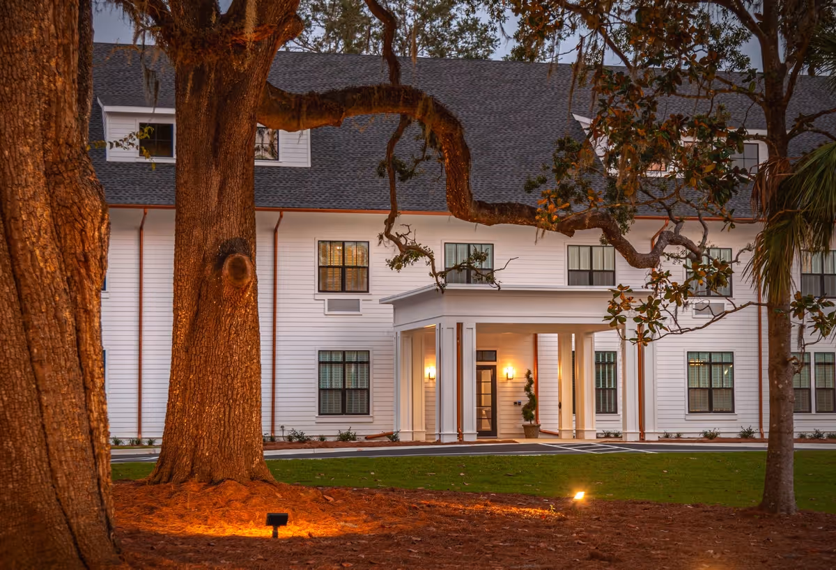 Exterior view of a large white assisted living facility building with multiple windows and a covered entrance, surrounded by large trees and a well-maintained lawn at dusk.