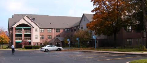 Three-story brick-and-stucco senior living building front with a parking lot, a white car, and trees with autumn foliage.