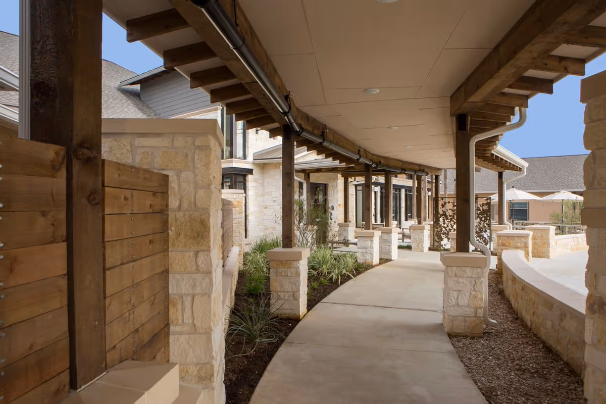 Covered stone and wood walkway along the exterior of a senior living building with columns and landscaping.