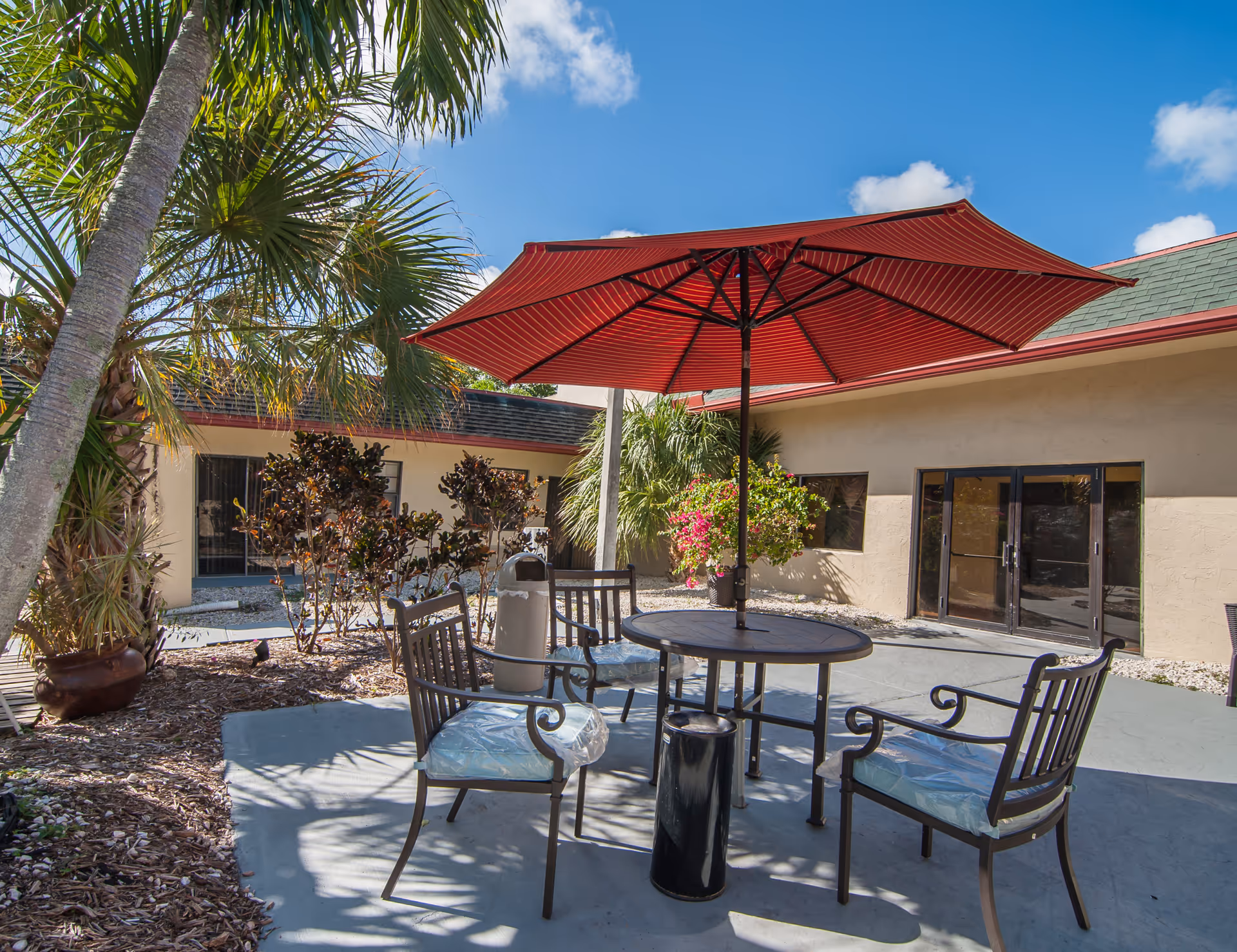 Outdoor patio area with a round table and four metal chairs with cushions under a large red umbrella. Surrounding the patio are palm trees, bushes, and a beige building with glass doors and windows under a clear blue sky.