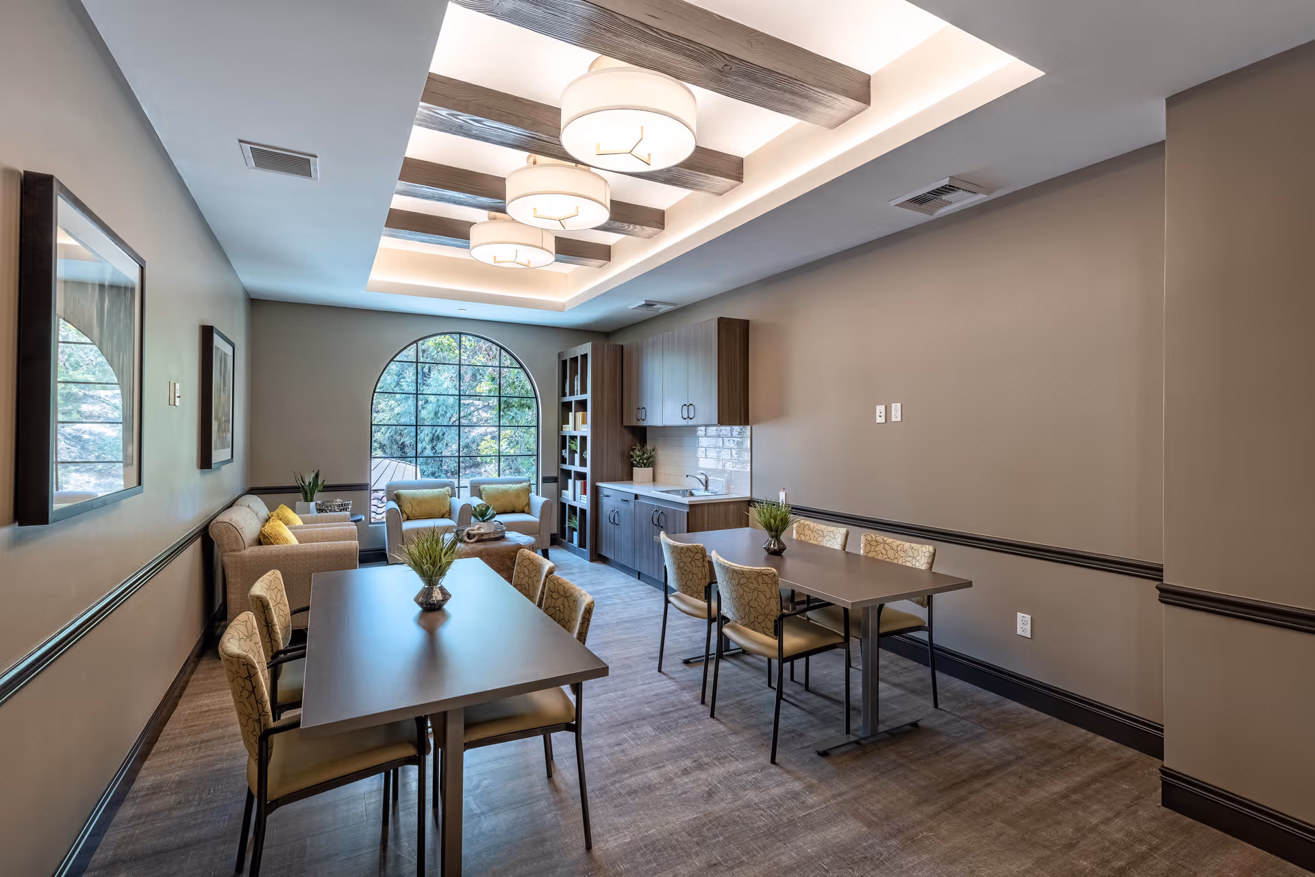 A well-lit common area with two rectangular tables surrounded by chairs, a small kitchenette with cabinets and a sink, and a seating area with two armchairs and a sofa near a large arched window showing greenery outside. The ceiling features exposed wooden beams and modern circular light fixtures.