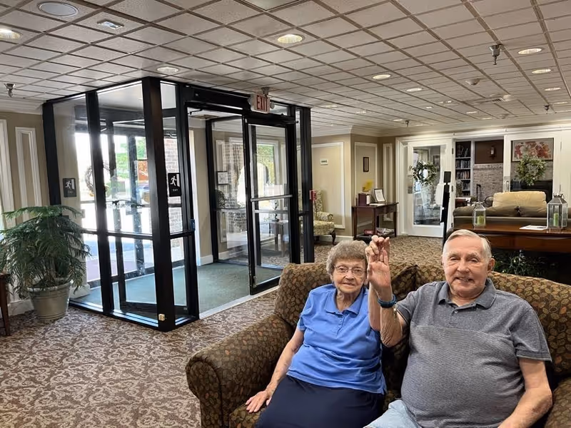 An elderly man and woman sitting on a patterned couch in a well-lit common area of a senior living facility. The man is holding the woman's hand up in a cheerful gesture. Behind them is a glass entrance door, potted plants, and a carpeted floor with a patterned design. The room has a cozy and welcoming atmosphere with additional seating and decorative elements.