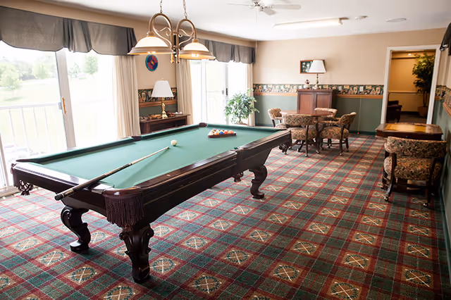 A recreational room with a green felt pool table in the center, a pool cue resting on it, and a rack of billiard balls arranged. The room has patterned carpet flooring, floral upholstered chairs around small tables, a lamp on a cabinet, large windows with curtains letting in natural light, and a ceiling fan.