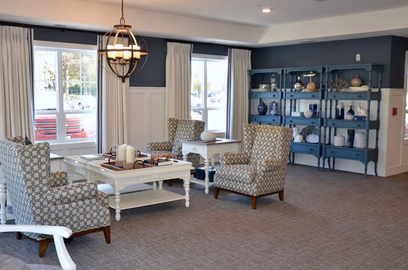 A cozy senior living common area with patterned armchairs arranged around white coffee and side tables. The room features large windows with white curtains, a blue shelving unit displaying decorative items, and a modern chandelier hanging from the ceiling. The carpeted floor and soft lighting create a welcoming atmosphere.