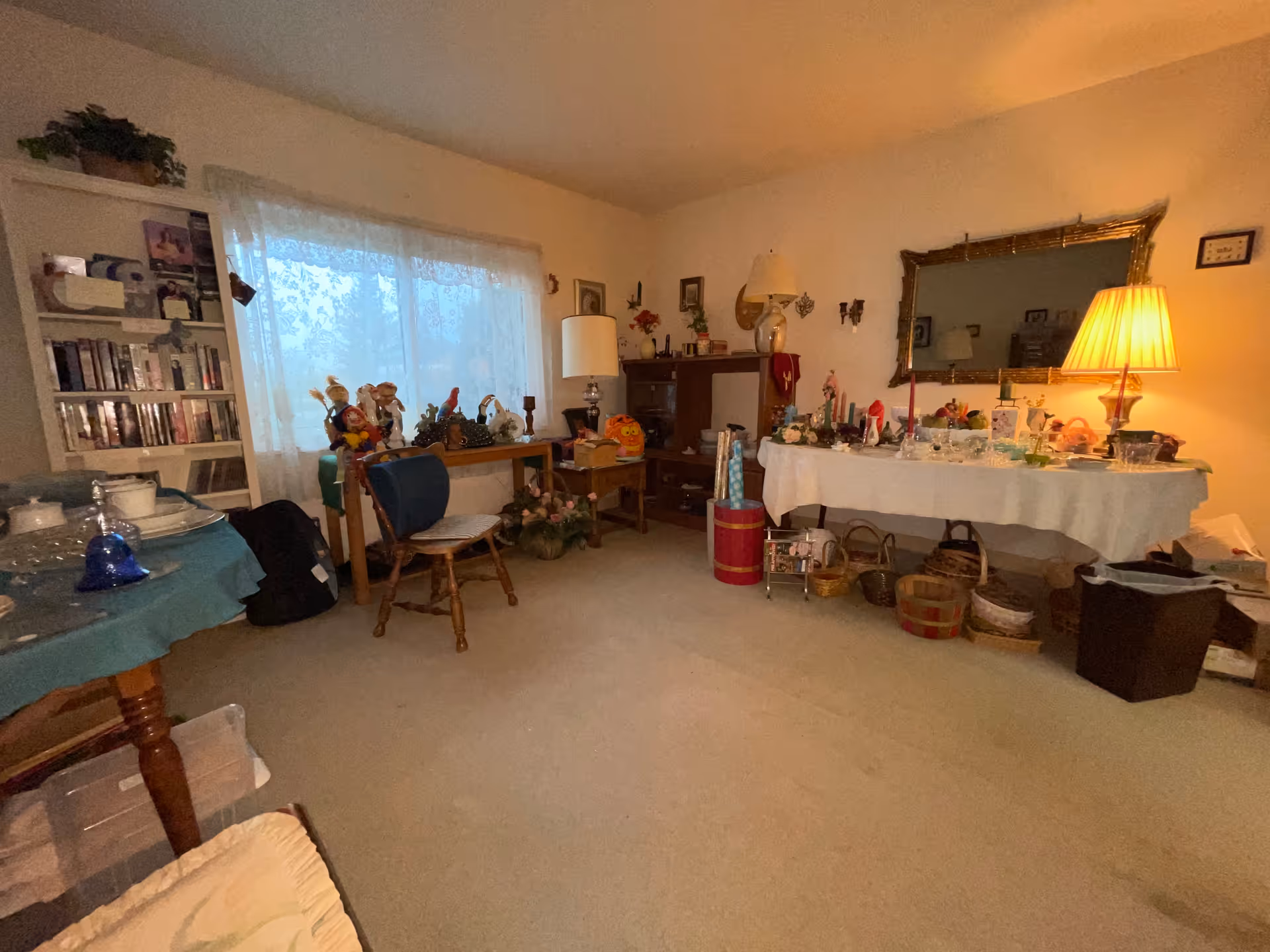 A cozy living room with beige carpet and cream-colored walls. There is a large window with sheer white curtains letting in natural light. The room contains various furniture including a wooden chair, a table covered with a white cloth displaying decorative items, a lamp with a beige shade, and a large ornate mirror on the wall. Shelves and tables hold various knick-knacks and decorations, creating a warm and lived-in atmosphere.