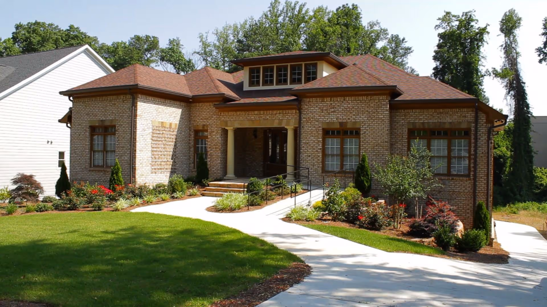 Front exterior view of a single-story brick building with a brown shingled roof, surrounded by a well-maintained lawn and landscaped garden beds with shrubs and flowers. A concrete walkway leads to the entrance, which has two columns supporting a small covered porch.