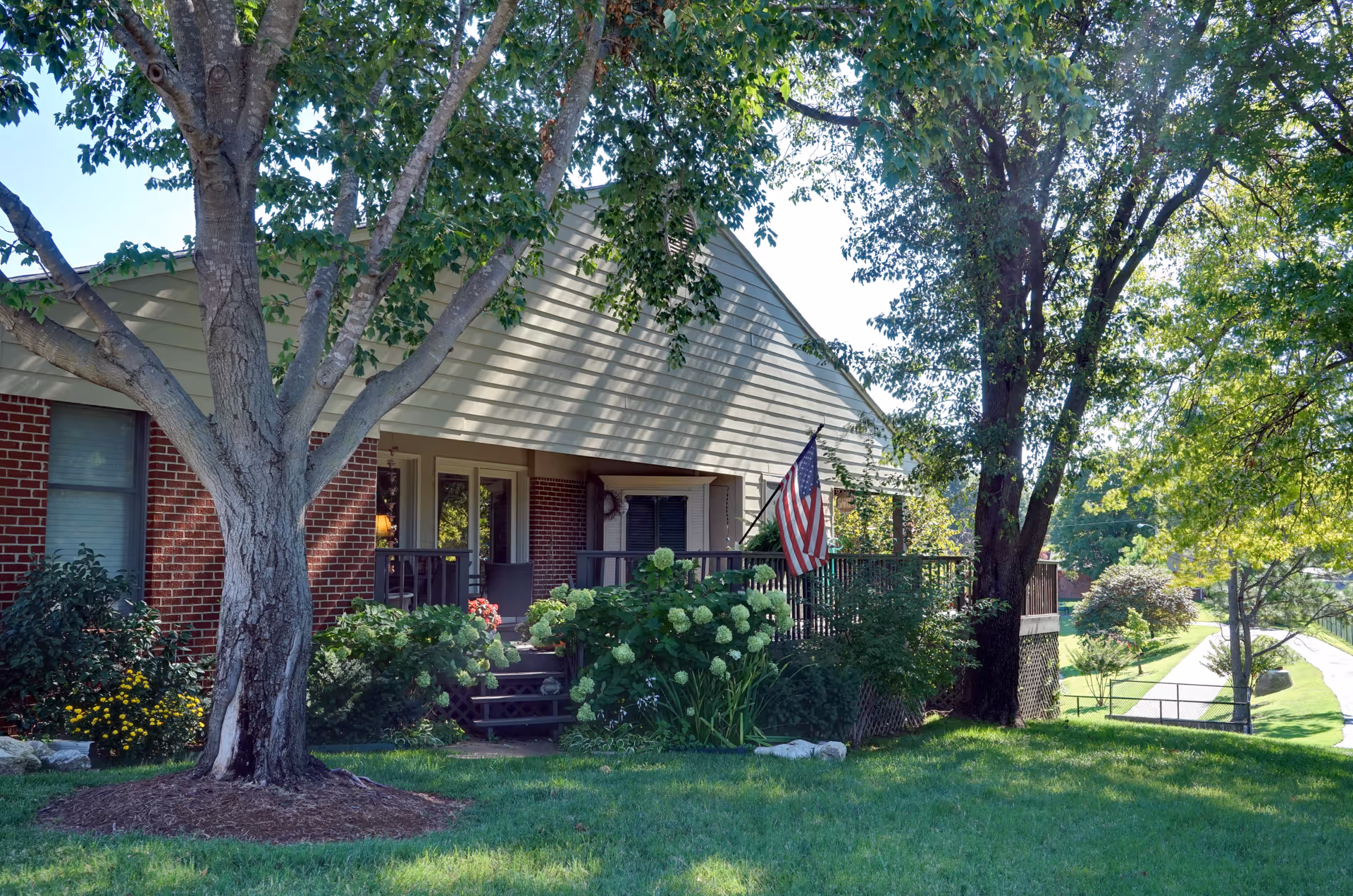 Exterior view of a single-story brick and siding building with a porch, surrounded by green trees and shrubs, with an American flag displayed on the porch.