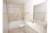 A clean and simple bathroom featuring a white bathtub with a shower curtain rod, a toilet, a sink with a countertop, a large mirror above the sink, and beige tiled flooring.
